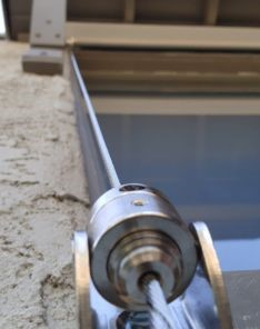 Close-up of metal hardware anchoring the tension cable for automated outdoor window shades on a textured stucco wall.